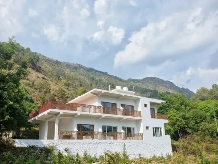 A white hill-side guesthouse with terraced balconies below a forested Kumaon ridge near Bhimtal