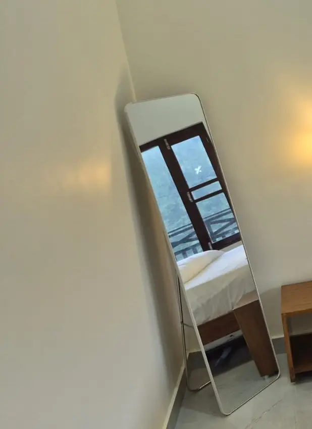 Full-length mirror in the corner of the room reflecting the window and bed